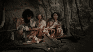 Ein Steinzeit Mann und zwei Frauen sitzen am Lagerfeuer in ihrer Höhle und braten sich Fleisch.